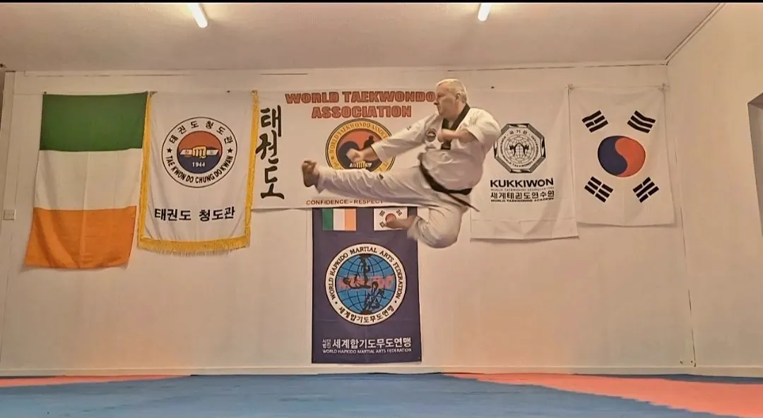 A World Taekwondo Ireland student mid-air in a flying kick in front of the Irish, association and Korean flags